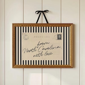 May include: A rectangular framed print featuring a vintage-style postcard design with the text 'from North Carolina, with love' in black script. The print is set against a black and white vertical striped background, encased in a gold frame with a black ribbon.