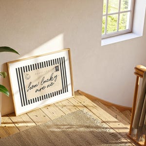 K&ouml;nnte beinhalten: Ein gerahmter Druck mit dem Text "how lucky are we" auf beigem Hintergrund mit schwarz-wei&szlig;en Streifen. Der Druck lehnt an einer Wand auf einer Holztreppe, mit einer Pflanze und einem Fenster im Hintergrund.