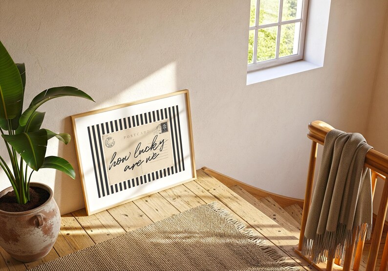 K&ouml;nnte beinhalten: Ein gerahmter Druck mit dem Text "how lucky are we" auf beigem Hintergrund mit schwarz-wei&szlig;en Streifen. Der Druck lehnt an einer Wand auf einer Holztreppe, mit einer Pflanze und einem Fenster im Hintergrund.
