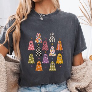 May include: A charcoal gray short-sleeve t-shirt featuring a graphic print of twelve stylized ghosts in various colors and patterns, including checkered, floral, leopard print, and wavy designs, arranged in a grid-like formation on the chest.