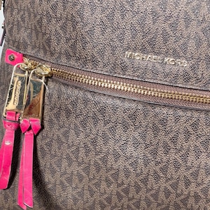Op de afbeelding: Bruine Michael Kors handtas met een herhalend MK-patroon. De tas heeft een gouden rits en gouden hardware met roze leren ritstrekkers. Het Michael Kors-logo is zichtbaar op de tas.