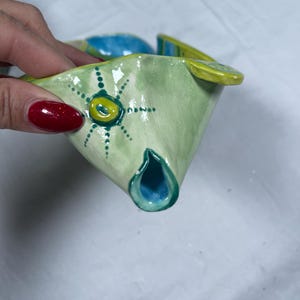 May include: A hand-painted ceramic fish-shaped bowl. The bowl features a light green body with yellow and blue accents. A sun-like design is painted on the side. The interior of the bowl is blue.