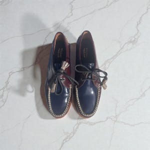May include: A pair of navy blue and burgundy leather boat shoes with white stitching. The shoes have a glossy finish and are tied with black laces and decorative tassels. The brand name "Weejuns" is visible on the inner lining.