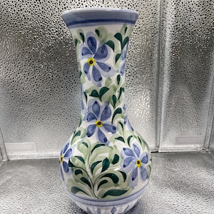 Może przedstawiać: Ceramiczny wazon z rozkloszowaną górą i zaokrągloną podstawą. Wazon pomalowany jest w niebieskie kwiaty, zielone liście i żółte środki na białym tle. Obręcz i podstawa mają niebieskie akcenty. Wazon ma około 25 cm wysokości.