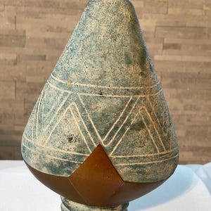 May include: A teal-colored, cone-shaped decorative object with a geometric pattern of white lines and triangles. The base is a dark brown color, and the bottom has a circular, ridged design. The object has a weathered, aged appearance.