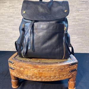 May include: Black leather backpack with a flap closure and a front zippered pocket. The backpack has a top handle and adjustable straps. The bag is sitting on a small, decorative wooden stool with carved details and brass accents.