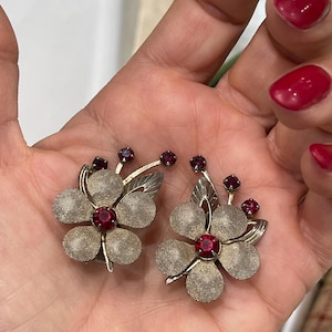 May include: A pair of vintage flower-shaped earrings. Each earring features a silver-toned mesh flower with a large, round, red gemstone center and smaller red gemstones. The earrings have silver-toned leaves.