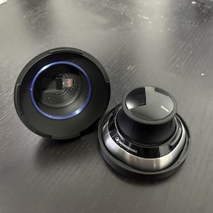 May include: A black and silver 3Dconnexion SpaceMouse wireless device, with a USB receiver and a black base. The device is open, showing the internal components. The device is on a dark surface.