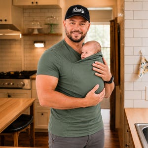 Op de afbeelding: Een saliegroen V-hals babydraagshirt met een ingebouwde buidel voor het dragen van een baby, gedragen met een zwarte honkbalpet met het woord Daddy in wit cursief geborduurd op de voorkant, gecombineerd met een grijze broek.