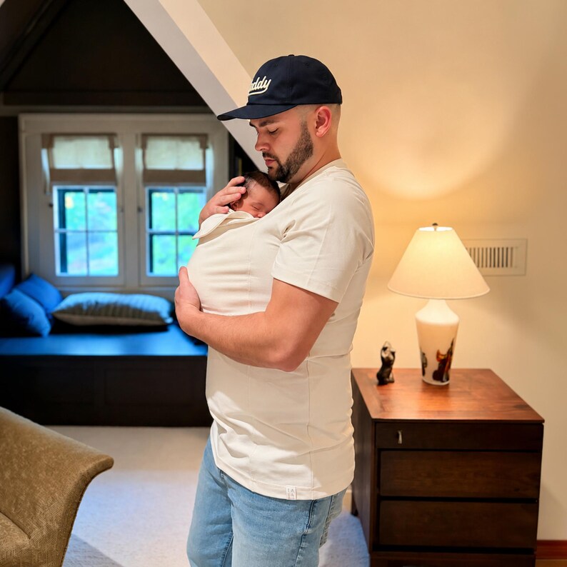 May include: A cream-colored short-sleeved kangaroo-style baby carrier shirt featuring a built-in pouch for holding an infant, paired with light blue denim jeans and a dark navy blue baseball cap with the word Daddy embroidered in white script.
