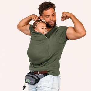 Op de afbeelding: Een olijfgroen T-shirt met korte mouwen in kangoeroestijl, voorzien van een ingebouwde buidel voor het dragen van een baby. Het shirt is ontworpen met een hoge halslijn en een ge&iuml;ntegreerde stoffen zak om een baby veilig tegen de borst te houden.