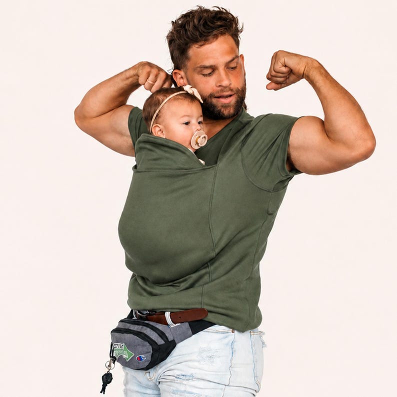 Op de afbeelding: Een olijfgroen T-shirt met korte mouwen in kangoeroestijl, voorzien van een ingebouwde buidel voor het dragen van een baby. Het shirt is ontworpen met een hoge halslijn en een ge&iuml;ntegreerde stoffen zak om een baby veilig tegen de borst te houden.