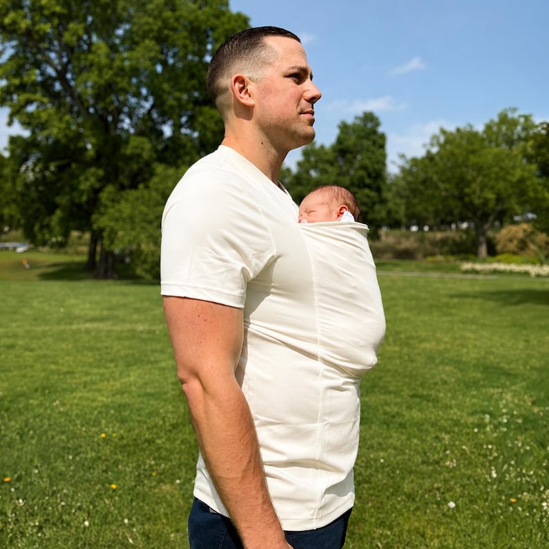 May include: A cream-colored short-sleeved shirt featuring an integrated fabric pouch on the front chest area, designed to hold a small infant securely against the torso. The garment is made of a smooth, stretchy material and has a fitted silhouette.