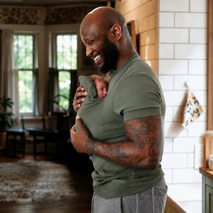 Op de afbeelding: Een olijfgroen baby-draagshirt met korte mouwen en een ingebouwde buidel op de borst voor het dragen van een baby. Het kledingstuk is gemaakt van eenkleurige stof en bevat een kapachtige flap die de bovenrug van de baby bedekt.