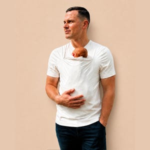 May include: A short-sleeved white t-shirt featuring an integrated fabric pouch on the chest area, designed to hold a small infant securely against the torso. The garment is worn with dark blue denim jeans, creating a functional and simple look.