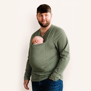 Op de afbeelding: Een olijfgroen shirt met lange mouwen en een V-hals, voorzien van een ge&iuml;ntegreerde stoffen buidel op de borst. In de buidel ligt een slapende baby, veilig gepositioneerd tegen het bovenlichaam. Het shirt wordt gedragen met een donkerblauwe spijkerbroek.