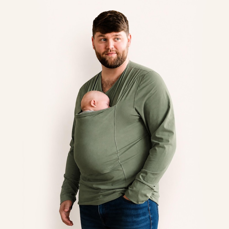 Op de afbeelding: Een olijfgroen shirt met lange mouwen en een V-hals, voorzien van een ge&iuml;ntegreerde stoffen buidel op de borst. In de buidel ligt een slapende baby, veilig gepositioneerd tegen het bovenlichaam. Het shirt wordt gedragen met een donkerblauwe spijkerbroek.