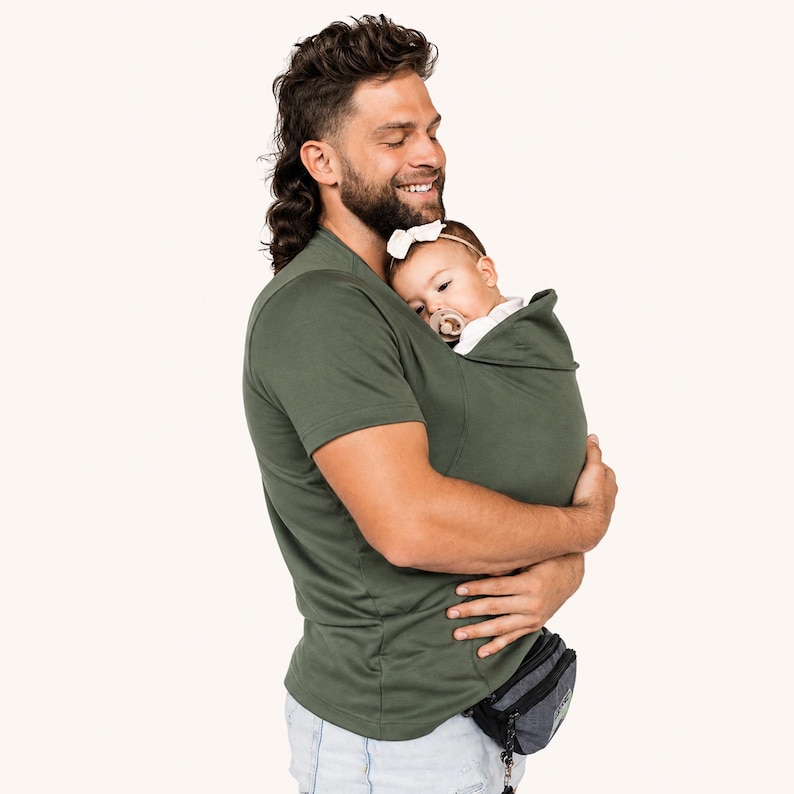 Op de afbeelding: Een olijfgroen baby-draagshirt met korte mouwen en een ingebouwde buidel voor het dragen van een baby. Het kledingstuk is gemaakt van een effen stof en bevat een kapachtige flap die het hoofdje van de baby ondersteunt terwijl deze tegen de borst rust.