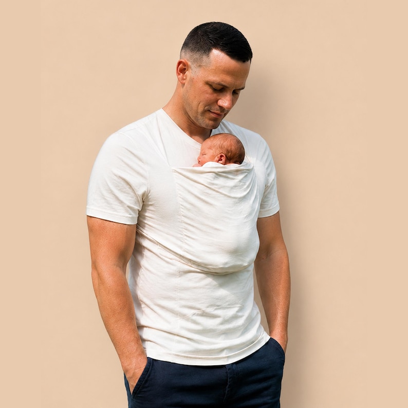 May include: A short-sleeved, off-white V-neck t-shirt featuring an integrated fabric pouch on the chest. A newborn infant is positioned inside the pouch, resting against the chest area of the shirt, which is worn with dark navy blue trousers.