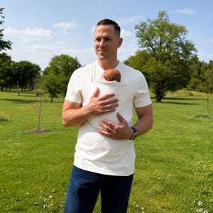 May include: A cream-colored short-sleeved shirt featuring an integrated fabric pouch on the chest designed to hold an infant. The shirt has a round neckline and a fitted silhouette, worn with dark blue denim jeans in an outdoor setting.