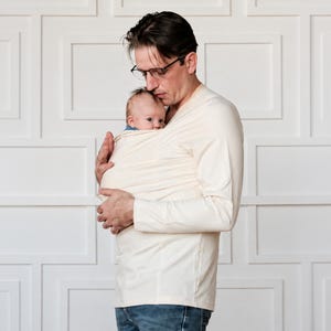 May include: A cream-colored long-sleeve shirt featuring an integrated fabric baby carrier pouch across the chest. The garment is made of a soft, solid-colored knit material with a V-neckline and long sleeves, designed to hold an infant securely.
