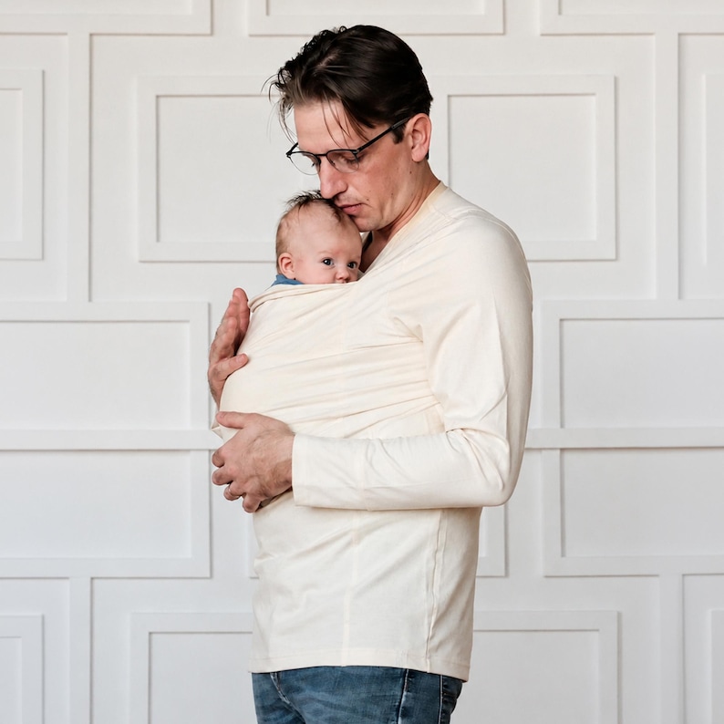 May include: A cream-colored long-sleeve shirt featuring an integrated fabric baby carrier pouch across the chest. The garment is made of a soft, solid-colored knit material with a V-neckline and long sleeves, designed to hold an infant securely.