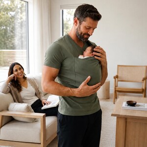 Op de afbeelding: Een olijfgroen babydraagshirt met V-hals en een ge&iuml;ntegreerde buidel op de borst waarin een slapende pasgeborene ligt. Het shirt is gemaakt van zachte stof en is ontworpen om de baby veilig en dicht tegen de borst aan te houden.