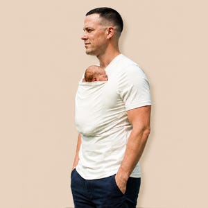 May include: A cream-colored short-sleeved t-shirt featuring an integrated fabric pouch on the chest area, designed to hold a small infant securely against the torso. The shirt is paired with dark navy blue trousers, creating a functional garment.
