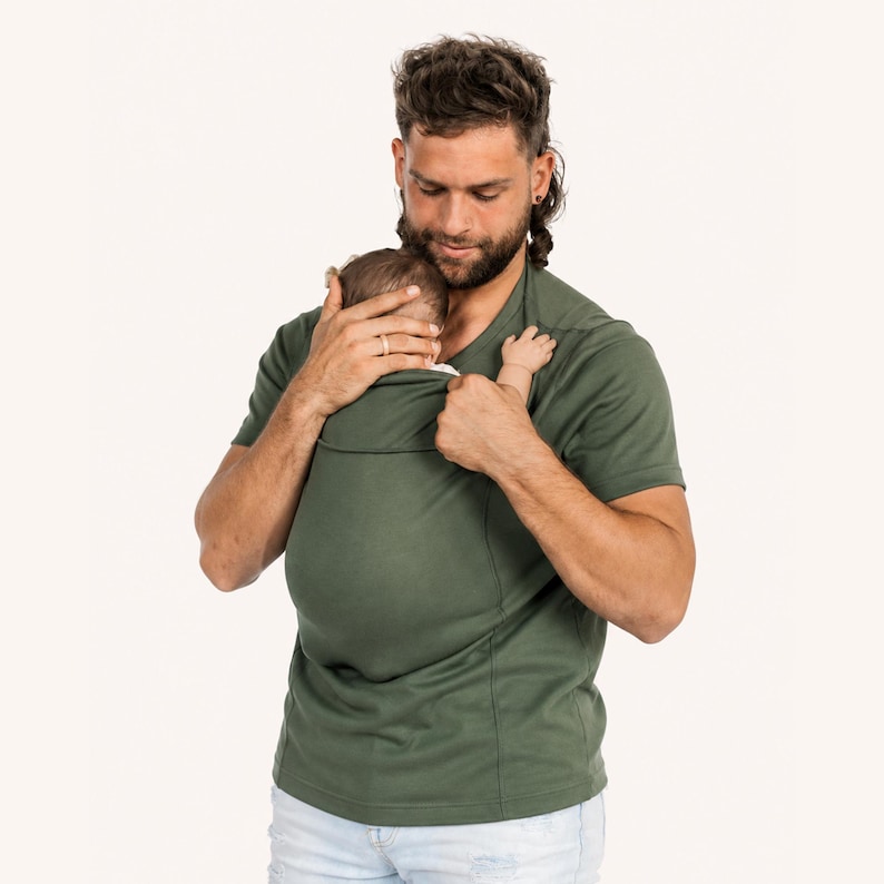 Op de afbeelding: Een effen olijfgroen T-shirt met korte mouwen en een ge&iuml;ntegreerde stoffen buidel op de borst, ontworpen om een baby in te dragen. Het kledingstuk heeft een ronde hals, een rechte zoom en is gemaakt van gladde, rekbare stof.