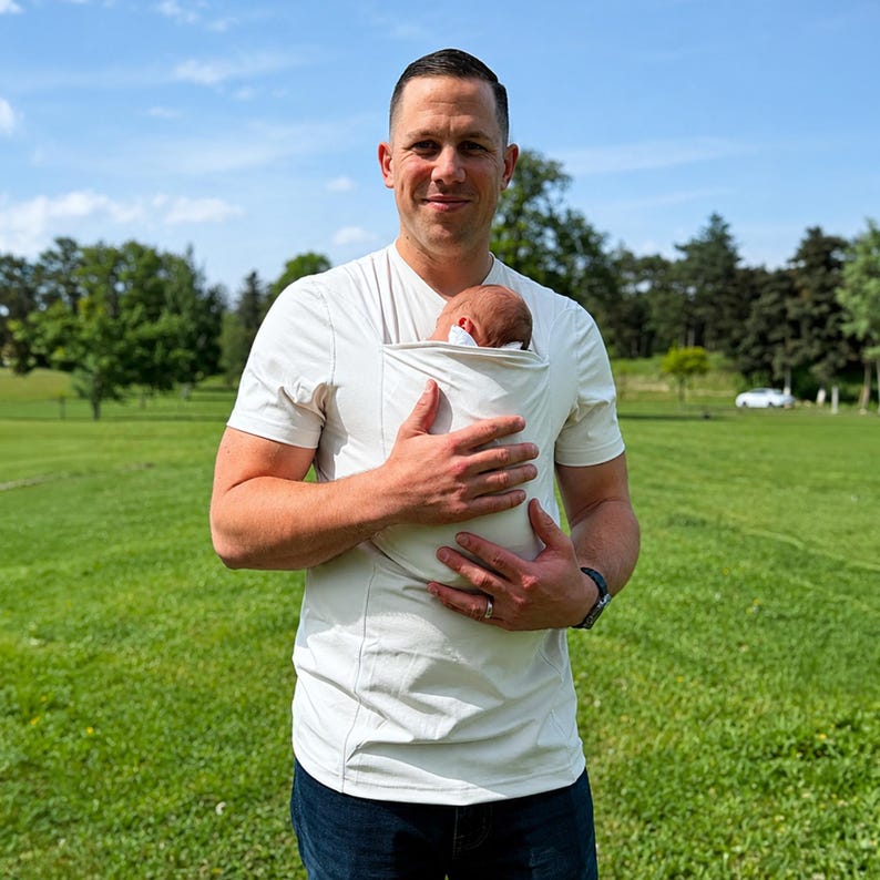 May include: A short-sleeved, off-white baby-wearing shirt featuring a built-in fabric pouch on the chest designed to hold an infant securely against the torso. The garment has a crew neckline and a fitted silhouette, constructed from a solid-colored fabric.