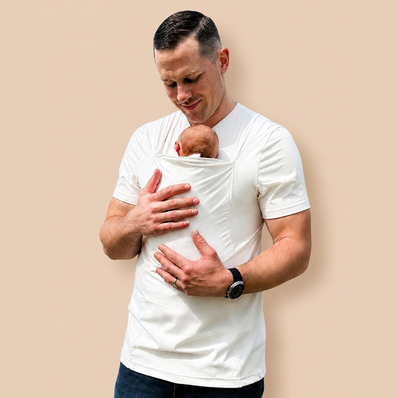May include: A short-sleeved, off-white crew neck t-shirt featuring an integrated fabric pouch on the front chest area designed to hold an infant securely against the torso. The garment is constructed from a smooth, lightweight, solid-colored fabric.