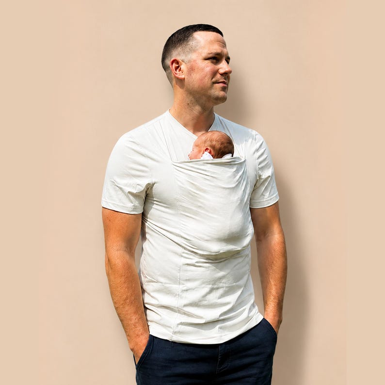 May include: A short-sleeved, off-white V-neck t-shirt featuring an integrated fabric pouch on the chest area. The pouch is designed to hold a small infant securely against the torso. The garment is paired with dark navy blue trousers.