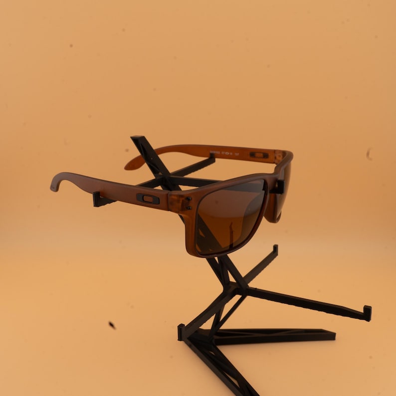 May include: Brown sunglasses with a rectangular frame and tinted lenses. The sunglasses are displayed on a black stand against a light orange background. The arms of the sunglasses are a lighter shade of brown.