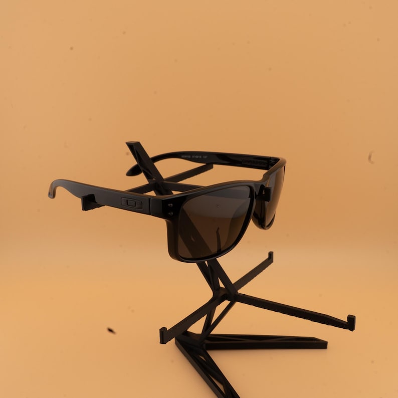 May include: Black sunglasses with dark lenses are displayed on a black stand. The sunglasses have a rectangular frame and are angled slightly. The stand has a geometric design with multiple support arms.