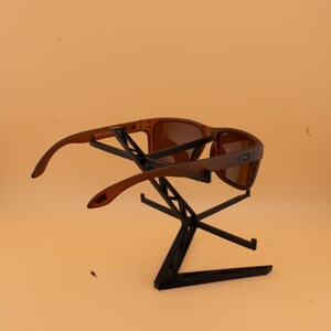 May include: Brown sunglasses with a rectangular frame are displayed on a black stand. The sunglasses have a matte finish and tinted lenses. The stand is made of black metal and has a geometric design.