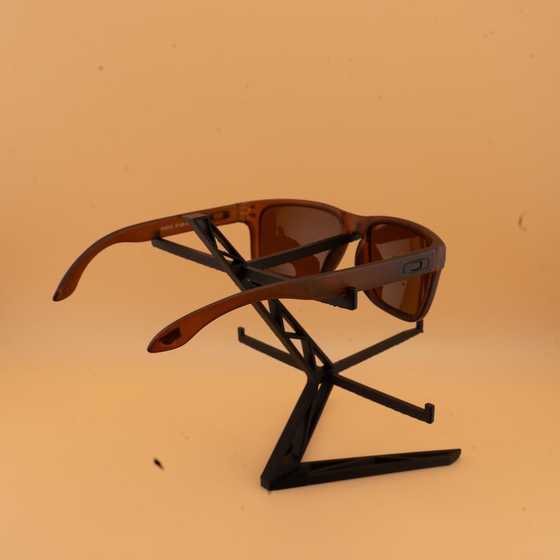 May include: Brown sunglasses with a rectangular frame are displayed on a black stand. The sunglasses have a matte finish and tinted lenses. The stand is made of black metal and has a geometric design.