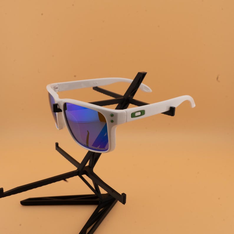 May include: White sunglasses with blue mirrored lenses. The sunglasses have a white frame with a green and black logo on the temple. The sunglasses are displayed on a black stand against a peach background.