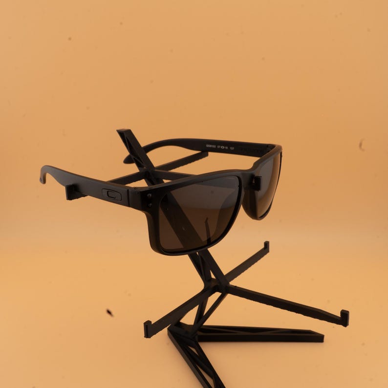 May include: A pair of black sunglasses with dark lenses, displayed on a black stand. The sunglasses have a rectangular frame and a modern design. The stand is black and has a unique, geometric shape, suitable for displaying eyewear.
