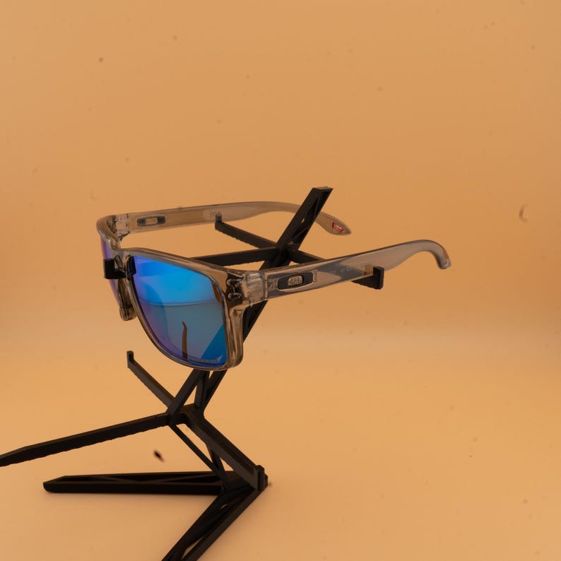 Puede incluir: Gafas de sol con montura transparente y lentes azules reflectantes. Las gafas de sol se exhiben en un soporte negro sobre un fondo naranja claro. Las monturas tienen forma rectangular.