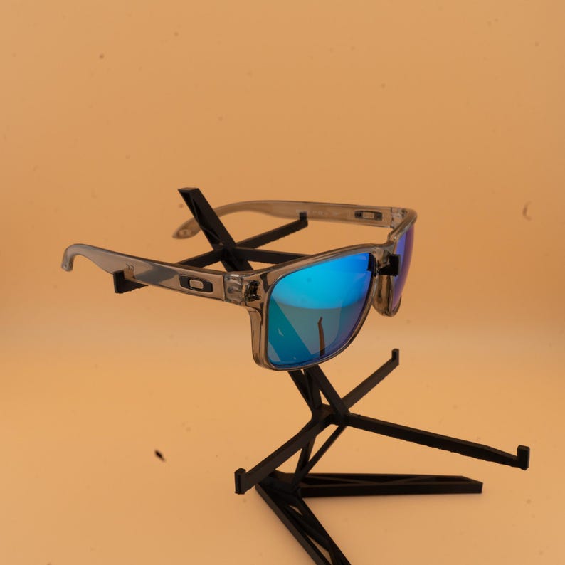 Puede incluir: Gafas de sol con montura transparente y lentes azules espejadas. Las gafas de sol se exhiben en un soporte negro sobre un fondo naranja claro. Las monturas son de color gris transl&uacute;cido.