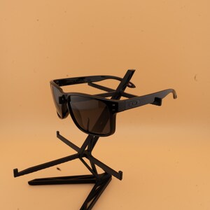 May include: Black sunglasses with dark lenses. The sunglasses have a square frame and are displayed on a black stand. The lenses appear to be tinted brown. The arms of the sunglasses are straight and have a small logo.