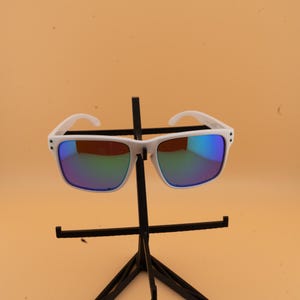 May include: White-framed sunglasses with square lenses and a reflective, multi-colored coating. The sunglasses are displayed on a black stand against a light orange background. The lenses reflect shades of blue, green, and yellow.