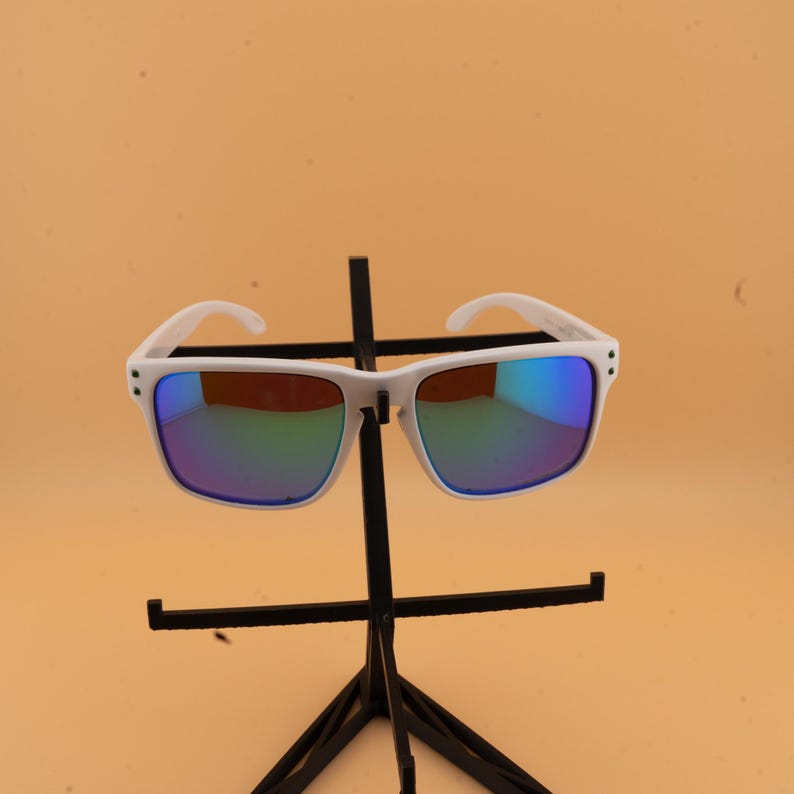 May include: White-framed sunglasses with square lenses and a reflective, multi-colored coating. The sunglasses are displayed on a black stand against a light orange background. The lenses reflect shades of blue, green, and yellow.