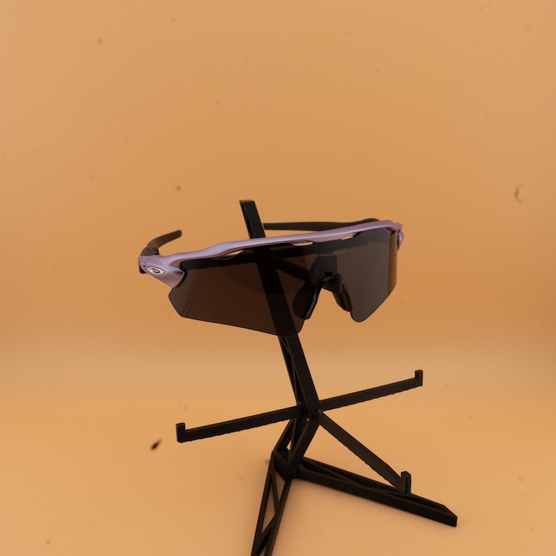 以下が含まれることがあります： ライトパープルのフレームと濃い色のレンズを備えたスポーツサングラス。黒いスタンドに展示され、背景はオレンジ色です。 スタイリッシュでモダンなデザインで、スポーツやアウトドア活動に適しています。