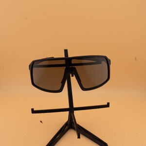 May include: Black sunglasses with brown lenses are displayed on a black stand. The sunglasses have a thick, rectangular frame. The stand is a tripod design, with a horizontal bar for the sunglasses to rest on.