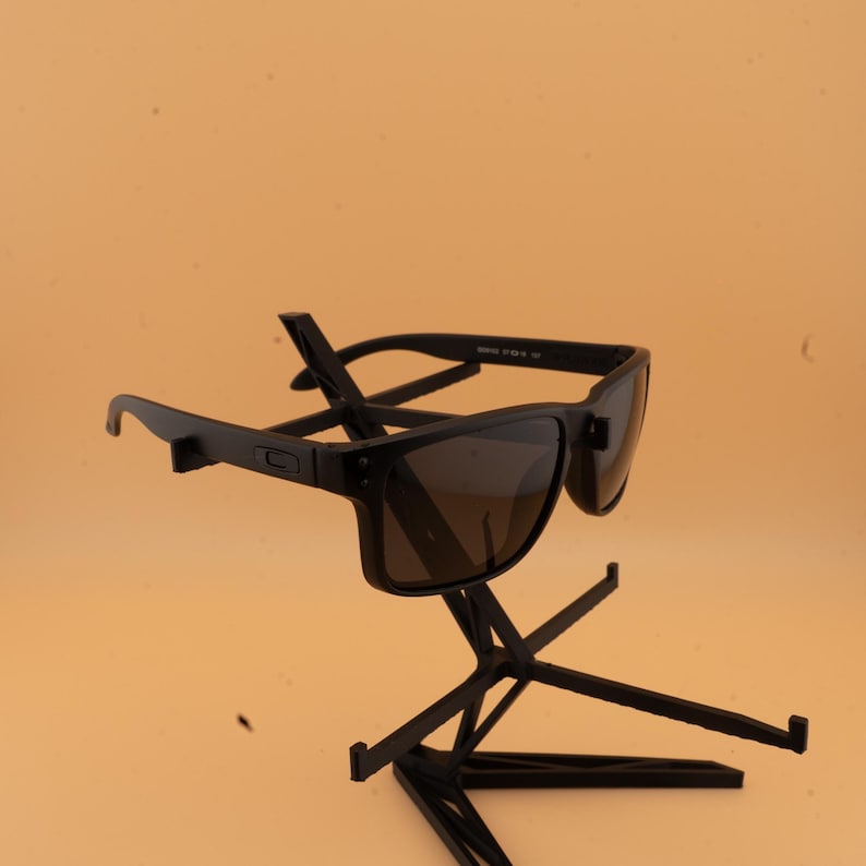 May include: A pair of black sunglasses with dark lenses, presented on a black stand against a light orange backdrop. The sunglasses feature a rectangular frame and a modern design. The stand is constructed from thin, intersecting black metal rods.