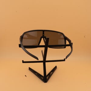 May include: Black sunglasses with a dark tint are displayed on a black metal stand. The sunglasses have a sporty design with a wide frame and curved temples. The stand has a geometric design.