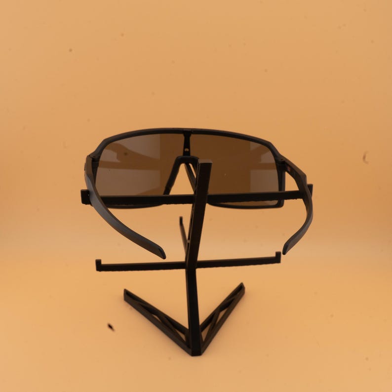 May include: Black sunglasses with a dark tint are displayed on a black metal stand. The sunglasses have a sporty design with a wide frame and curved temples. The stand has a geometric design.