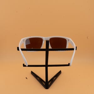 Puede incluir: Gafas de sol blancas con lentes marrones se exhiben en un soporte negro. Las gafas tienen una montura rectangular y un dise&ntilde;o deportivo. El soporte es de metal negro y tiene una forma geom&eacute;trica.