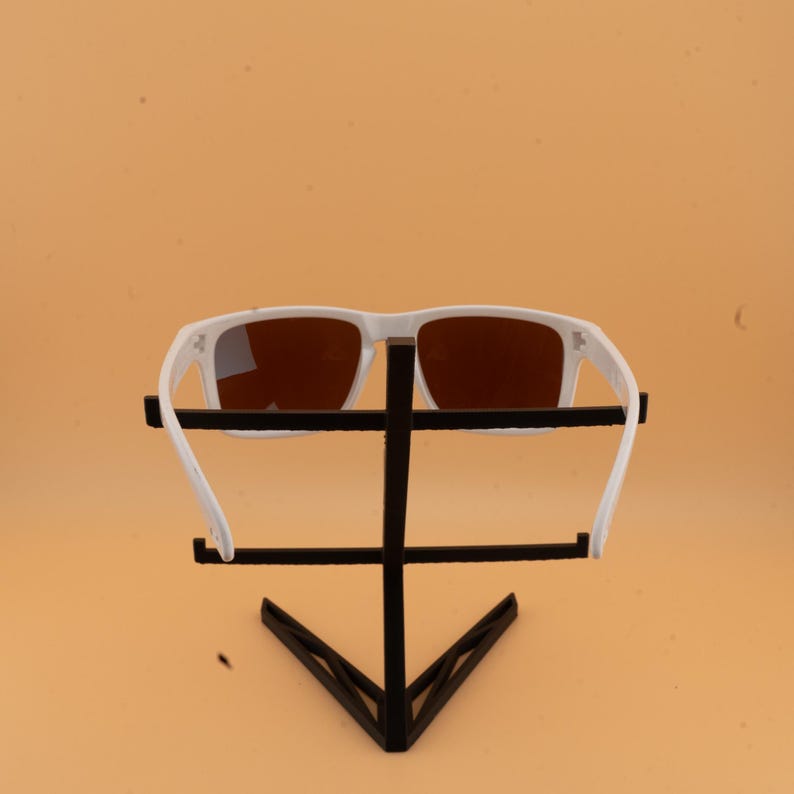 Puede incluir: Gafas de sol blancas con lentes marrones se exhiben en un soporte negro. Las gafas tienen una montura rectangular y un dise&ntilde;o deportivo. El soporte es de metal negro y tiene una forma geom&eacute;trica.